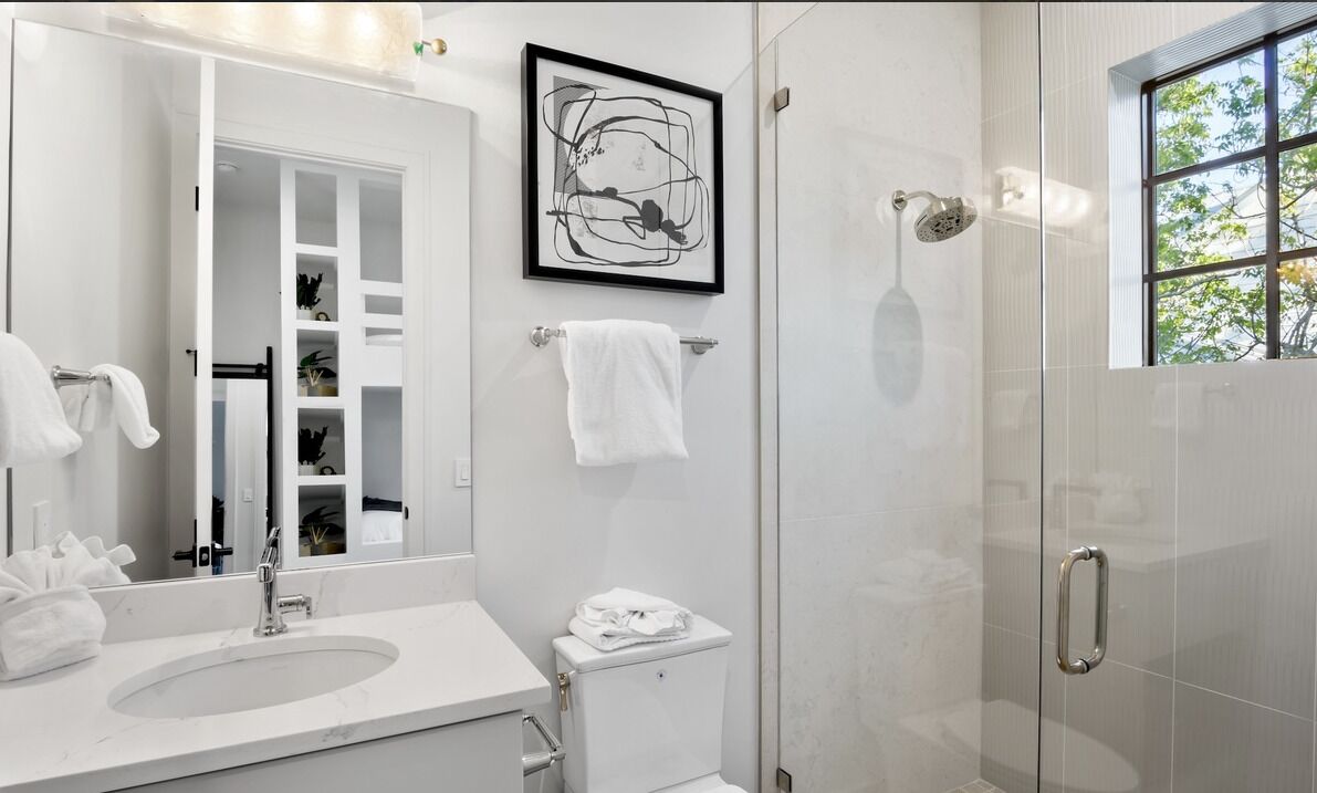 Een moderne badkamer met een grote spiegel, een wastafel, toilet en douche. Op de muur hangt een abstract kunstwerk in een lijst en een raam zorgt voor natuurlijk licht.