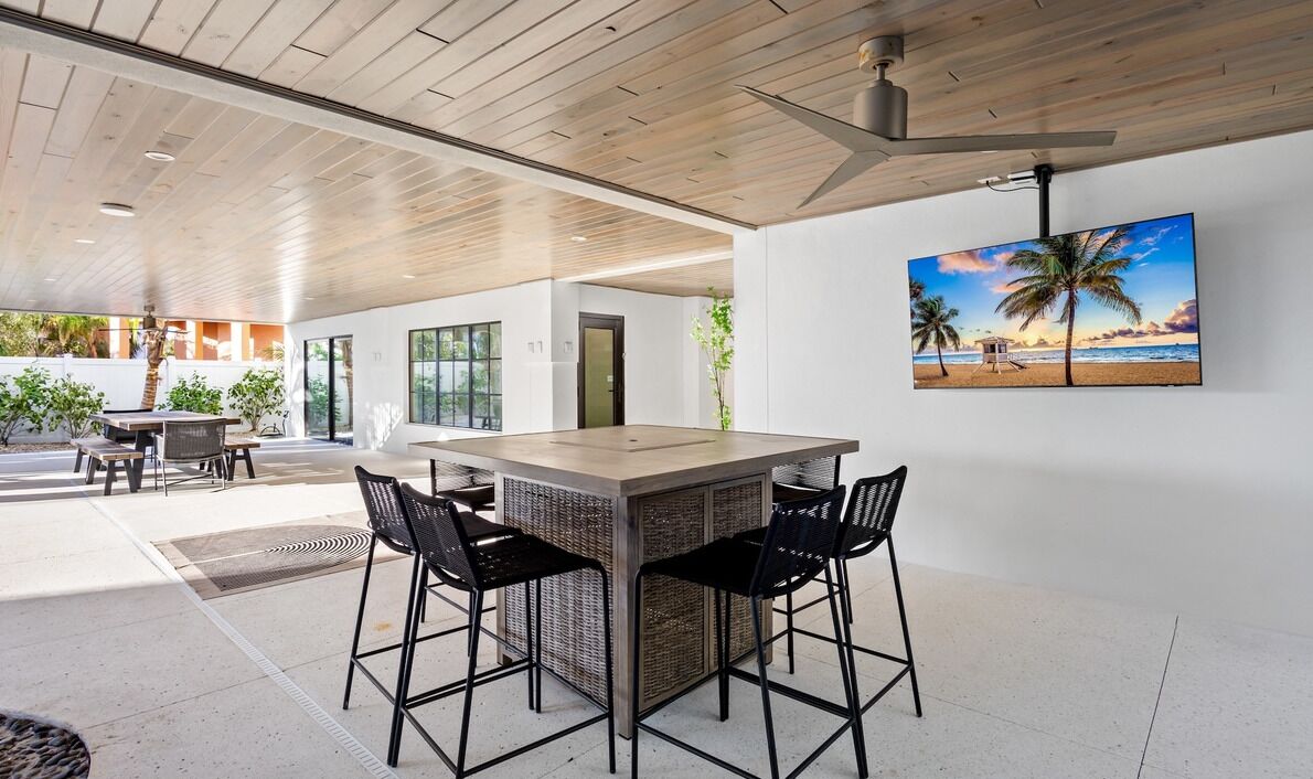 An outdoor bar area with a wooden ceiling, a large TV screen displaying a beach scene, and a seating arrangement with four chairs.