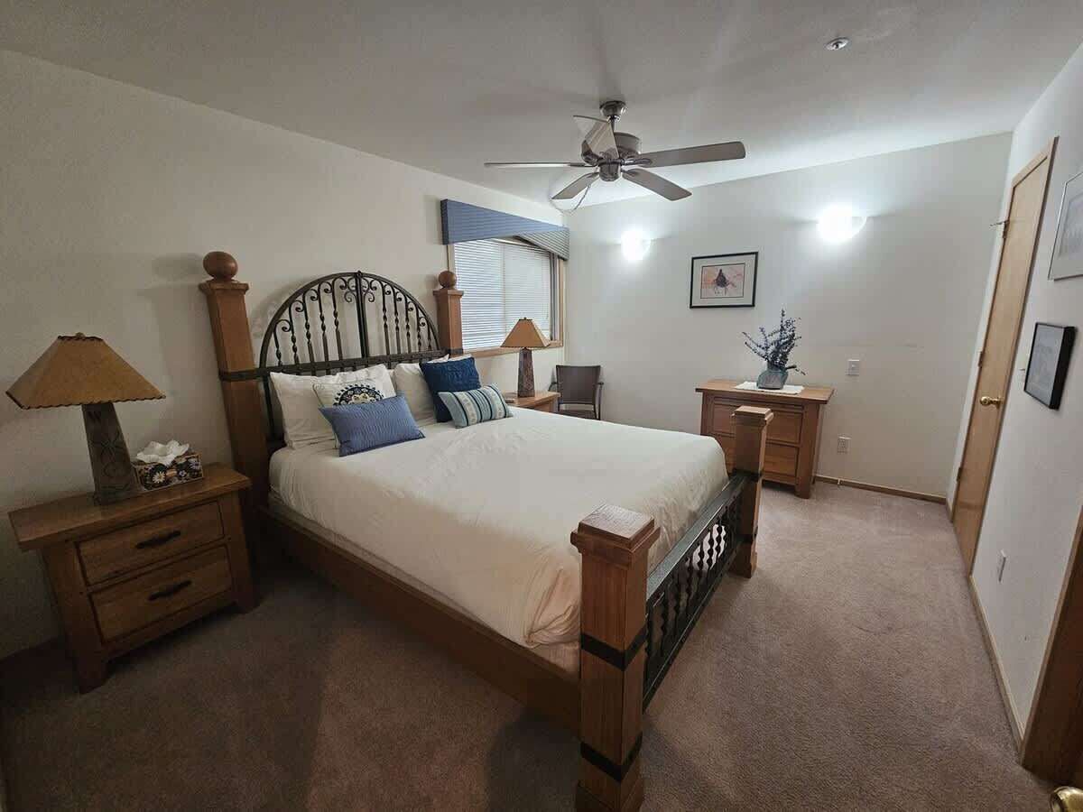 Un dormitorio con cama, mesitas de noche, escritorio, silla, ventilador de techo, ventana con persianas y puerta.