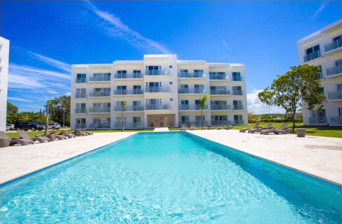 Um hotel moderno com piscina central, várias varandas e um céu azul límpido.