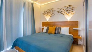 Un dormitorio con cabecera de madera, una cama con cubrecama azul, dos lámparas de pared y un ventanal con cortinas.