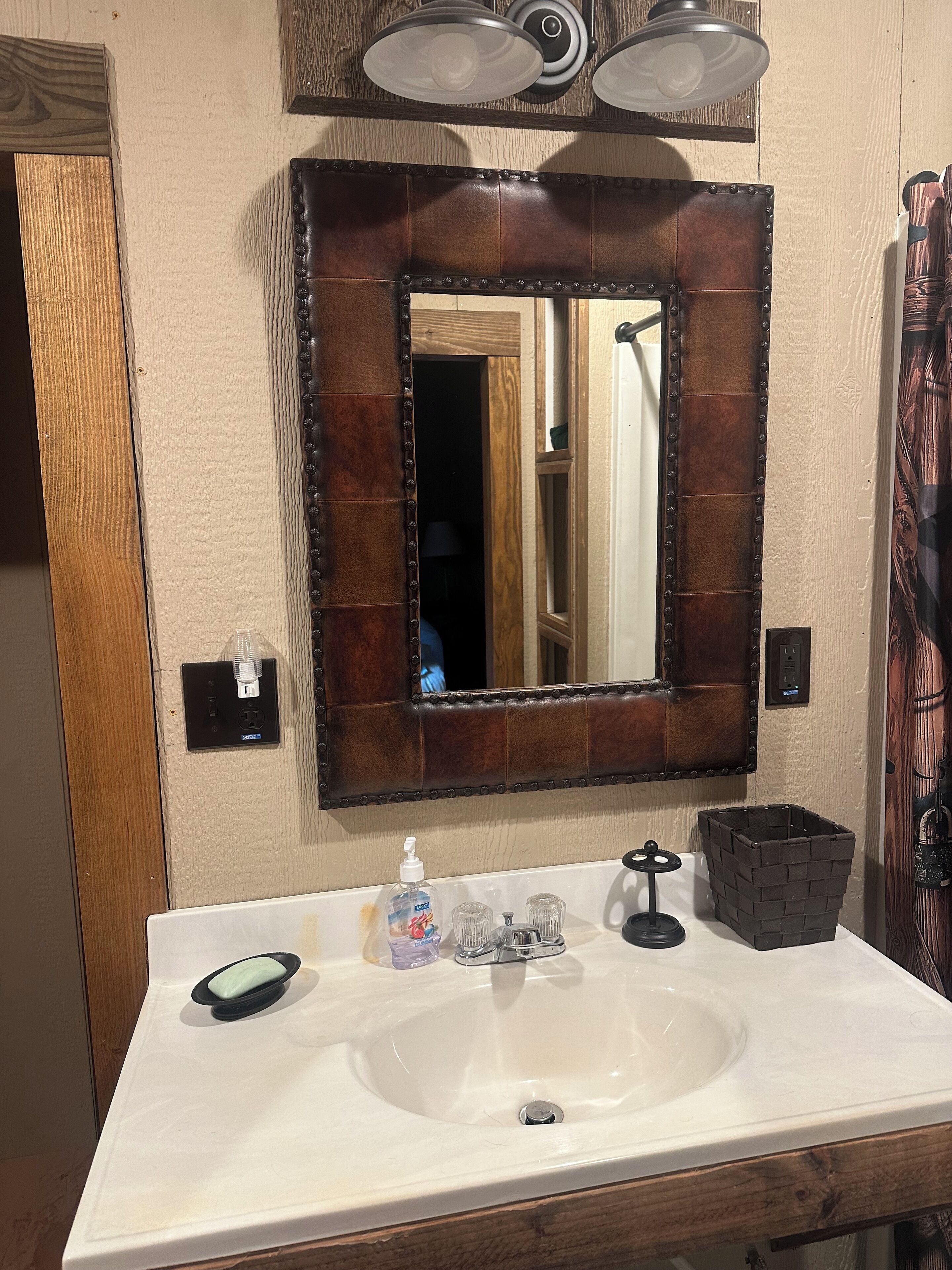 Un baño con un lavabo blanco, un espejo con marco de madera, una lámpara sobre el espejo y un cubo de basura negro.