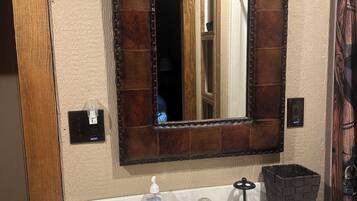 Un baño con un lavabo blanco, un espejo con marco de madera, una lámpara sobre el espejo y un cubo de basura negro.