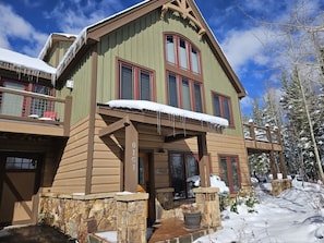 A two-story wooden house with a stone foundation, a balcony, and a snow-covered roof.