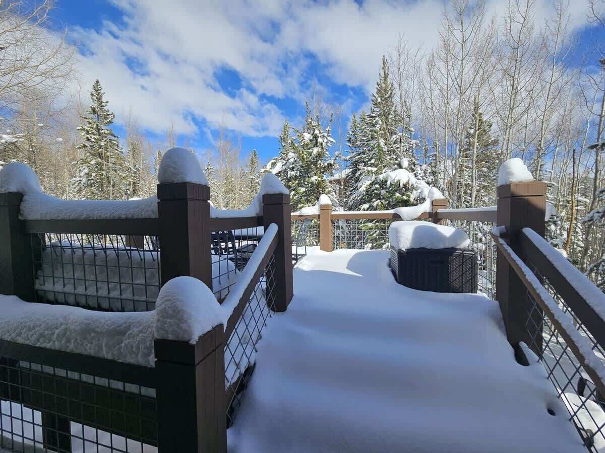 A snow-covered outdoor area with a metal fence and wooden posts, surrounded by trees and a clear sky.