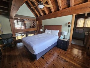 A bedroom with a wooden ceiling, a bed with white linens, a nightstand, a desk with a chair, and a window with a view of the outside.