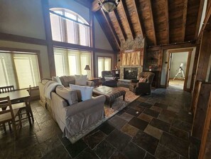A spacious living room with a high ceiling, large windows, a fireplace, and a stone-tiled floor.