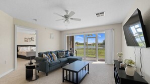 A spacious living room with a large sectional sofa, a coffee table, and a flat-screen TV. There is a ceiling fan and a sliding glass door leading to a balcony with a view of a park and lake.