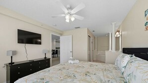 A bedroom with a bed, a TV, a ceiling fan, a dresser, and a bathroom visible through an open door.