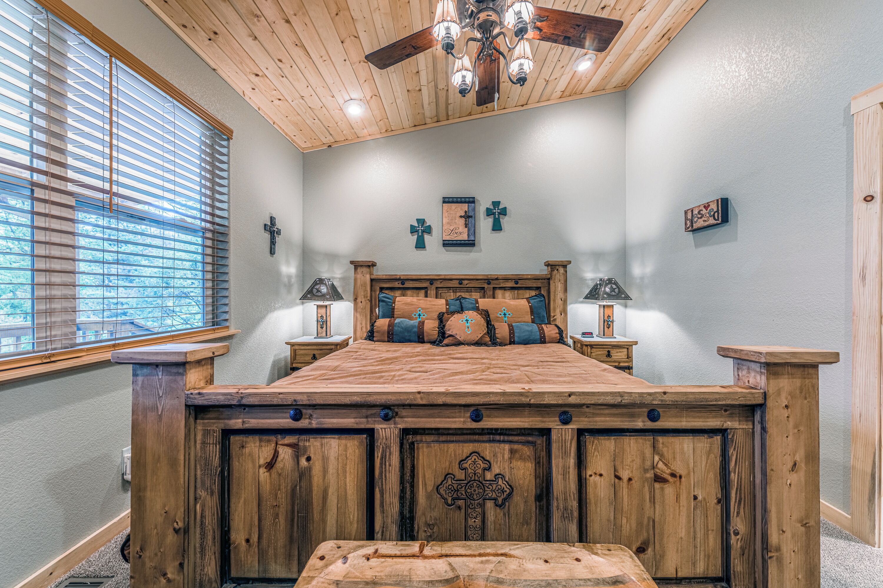 A bedroom with a wooden bed, two bedside tables with lamps, a cross on the wall, and a ceiling fan.