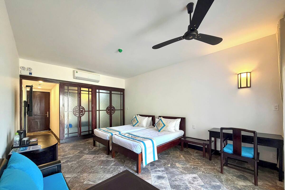 Une chambre d’hôtel comprenant un lit, un bureau, une chaise, un ventilateur de plafond et une porte avec des panneaux en verre dépoli.