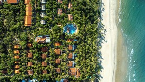 Eine Vogelperspektive zeigt ein Resort mit mehreren bungalowsartigen Unterkünften, umgeben von üppiger Vegetation, einem zentralen Pool und einem Strand neben der Anlage.