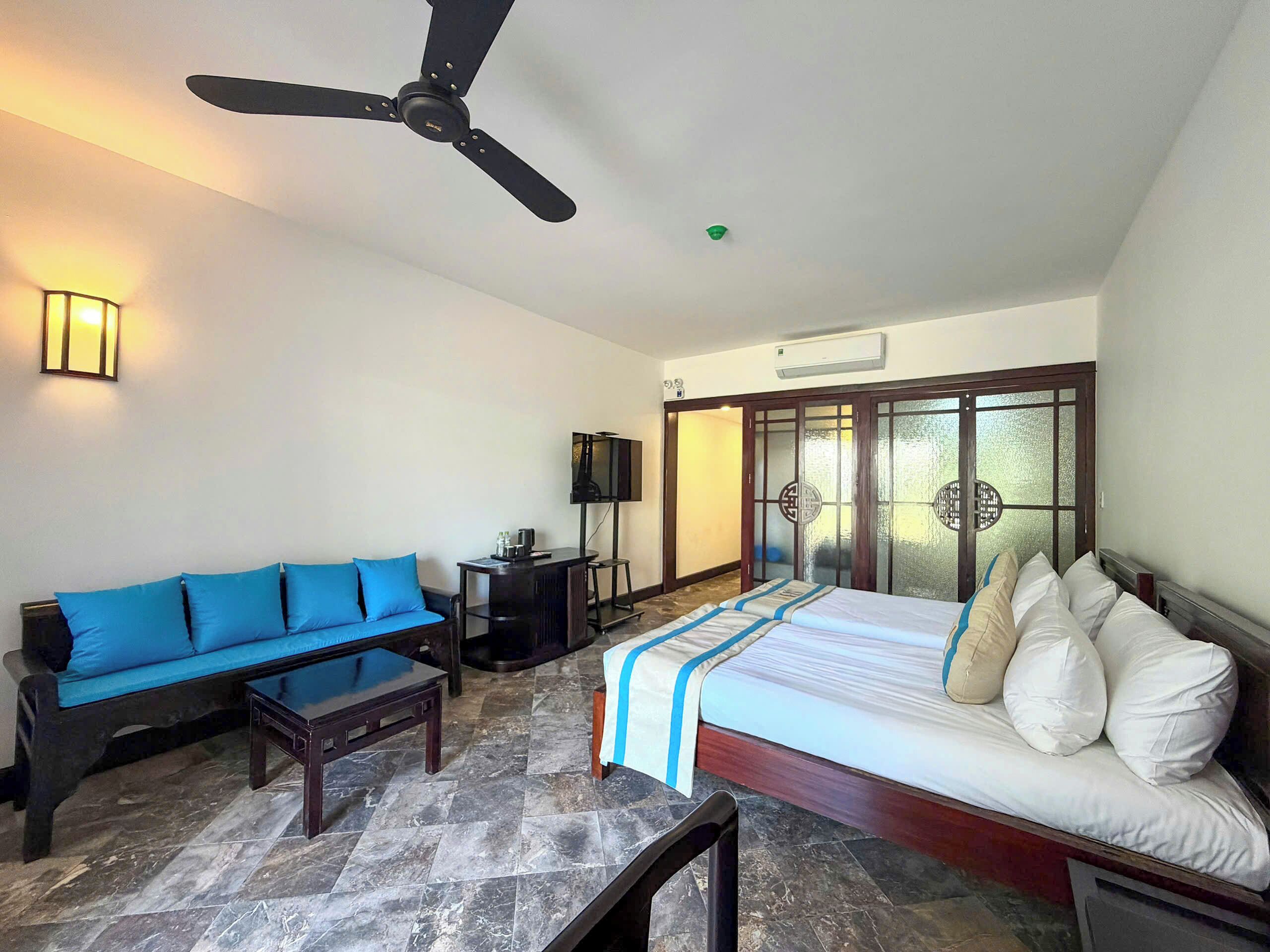 A hotel room with a bed, a blue sofa, a small table, a ceiling fan, a TV, and a sliding glass door.