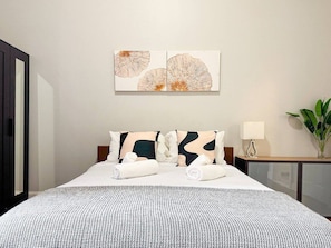 Een hotelkamer met een bed, twee kussens, een nachtkastje met een lamp en een kunstwerk aan de muur.