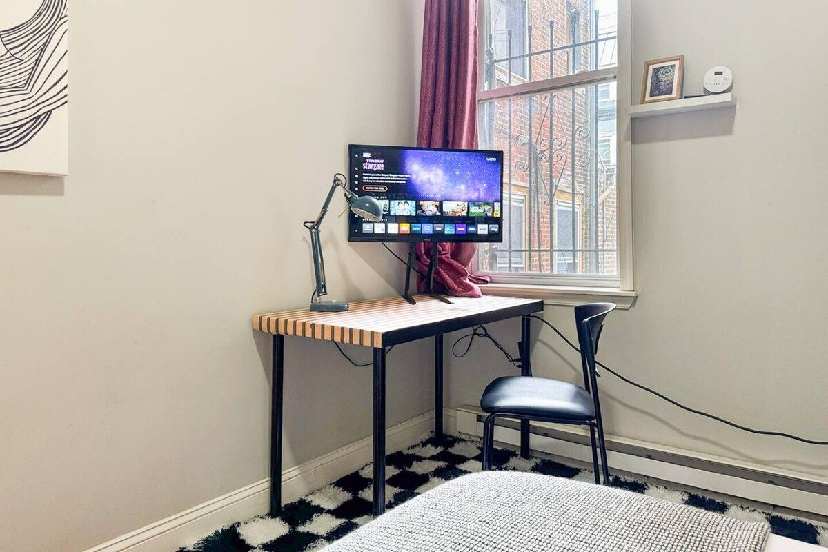 A room with a desk, a computer monitor, a chair, a window with curtains, and a wall-mounted shelf with a picture and a device.