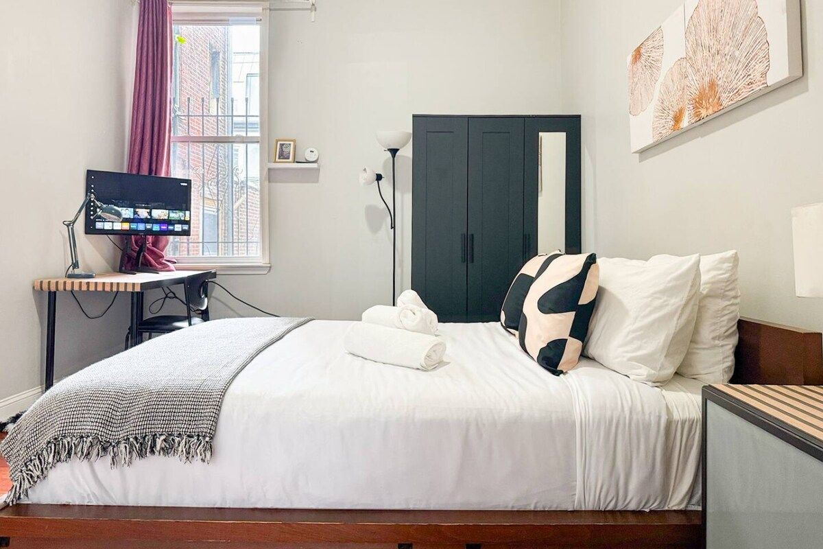 A neatly made bed with white bedding and a black and white pillow, a desk with a computer monitor, a lamp, and a window with curtains.