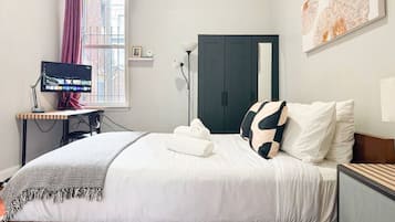 A neatly made bed with white bedding and a black and white pillow, a desk with a computer monitor, a lamp, and a window with curtains.