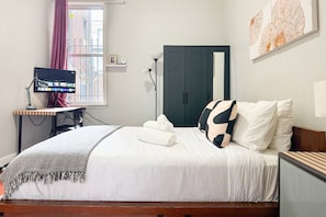 Un lit bien fait, avec une literie blanche et un coussin noir et blanc, un bureau avec un écran d’ordinateur, une lampe et une fenêtre avec des rideaux.