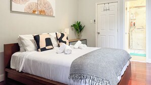 Un lit bien fait, avec du linge de lit blanc et un plaid à carreaux, une table de chevet ornée d’un vase contenant des plantes vertes, et une porte donnant sur une salle de bain où l’on aperçoit une douche et des toilettes.