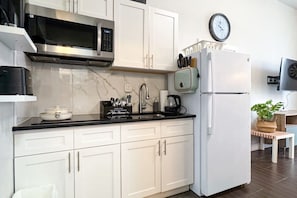 Een moderne keuken met witte kastjes, een RVS magnetron, een witte koelkast en een klok aan de muur.