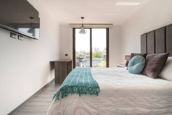 Quarto com cama, mesa de cabeceira, luminária, janela com vista da cidade e luz no teto.