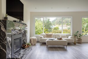 Un salon moderne avec une cheminée, un canapé blanc et de grandes fenêtres donnant sur un paysage verdoyant.
