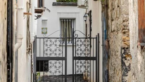 Une ruelle étroite, fermée par un portail, bordée de murs en pierre et dotée d’une petite fenêtre donnant sur la rue.