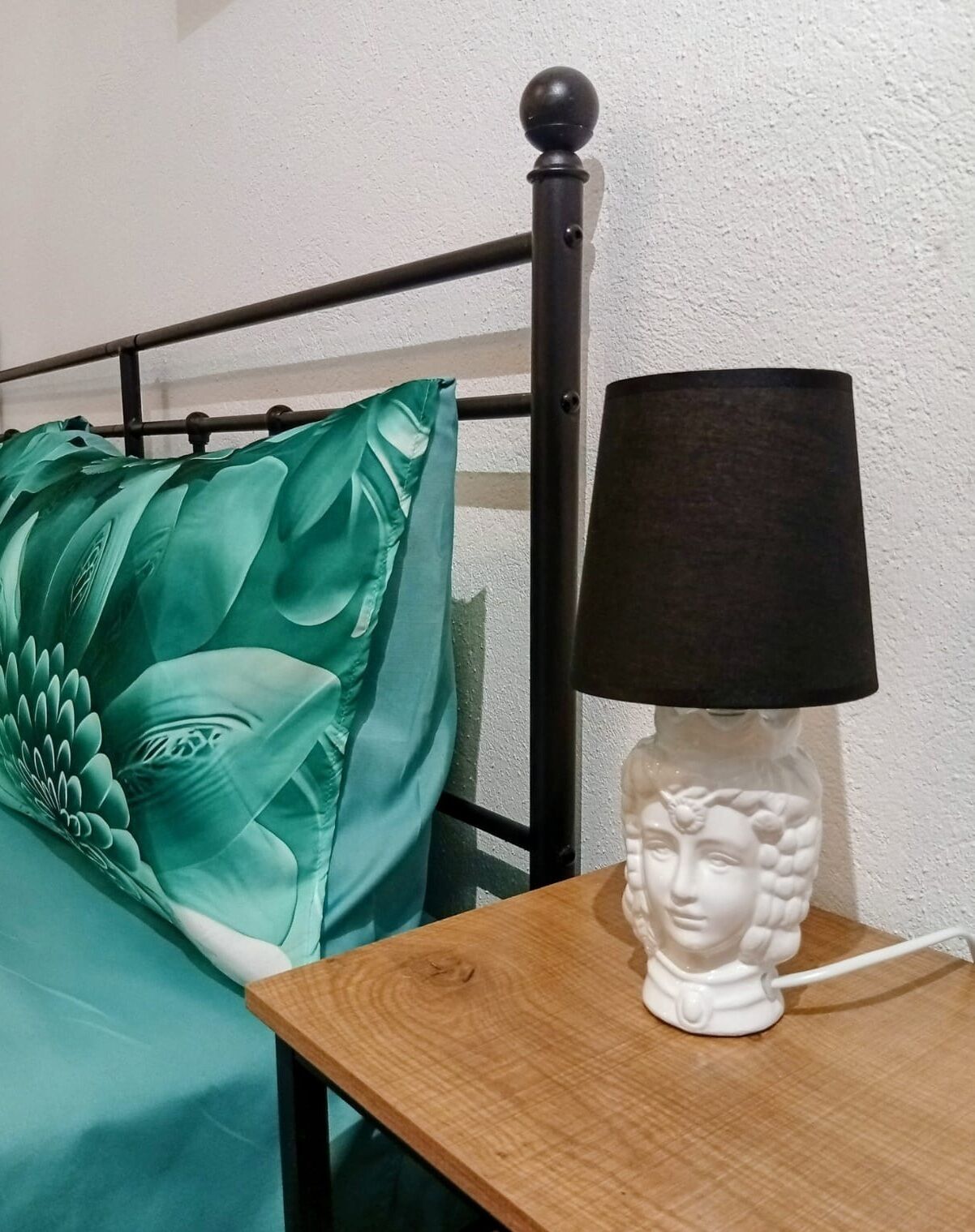 Ein Bett mit einem grünen Kopfkissen und einer weißen Lampe mit schwarzem Lampenschirm auf einem Nachttisch aus Holz.