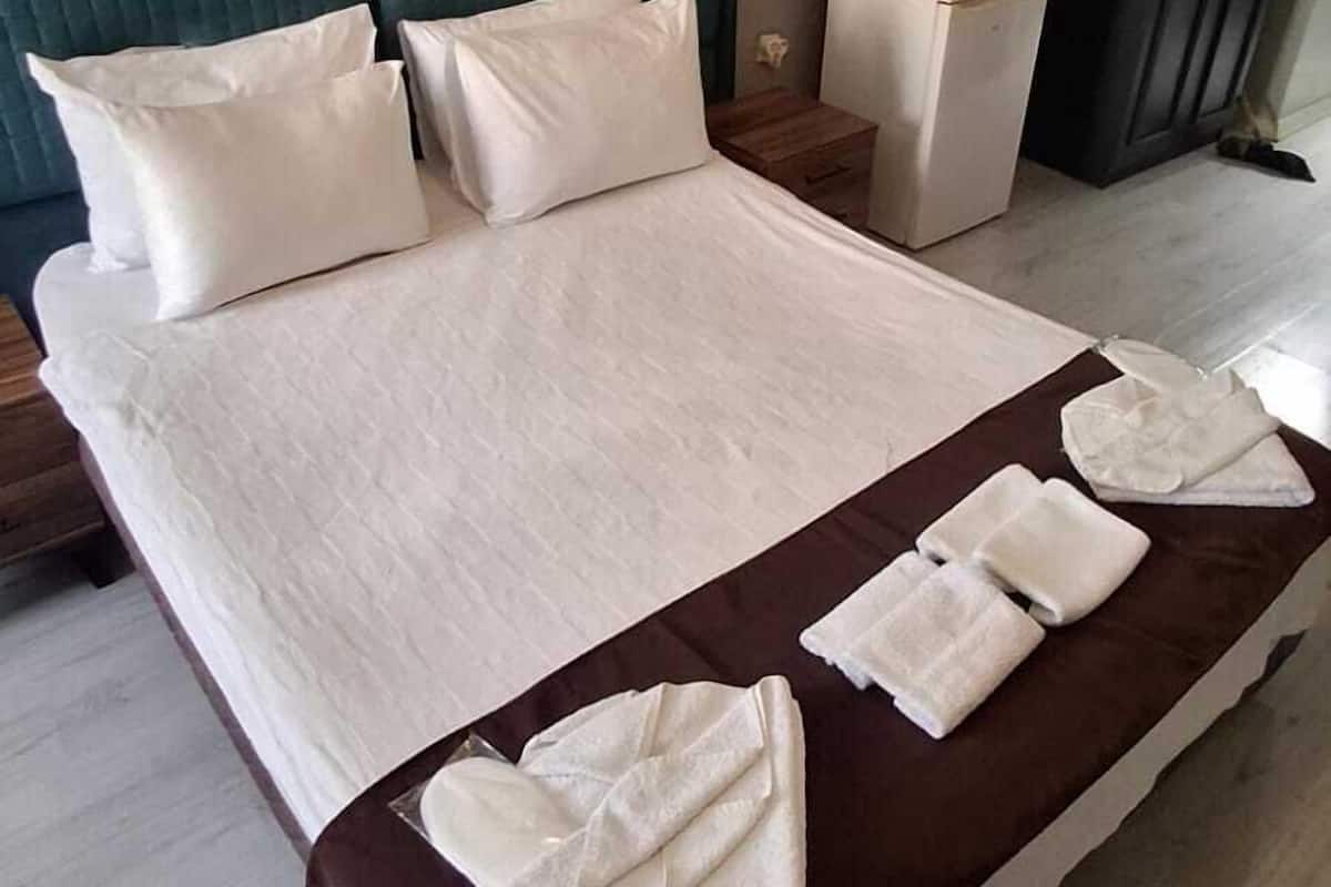 A neatly made bed with white linens and a brown accent, a nightstand with a kettle, a painting of a coastal scene, and a dark wardrobe.