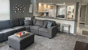 Una sala moderna con un sofá seccional gris, un pequeño reposapiés y una mesa de centro. Se observa un ventilador de techo e iluminación empotrada.