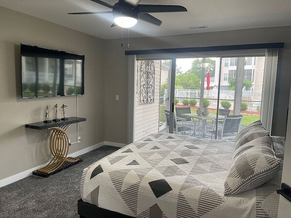 A bedroom with a bed, a TV mounted on the wall, a ceiling fan, and a view of the outside.