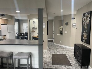 A modern kitchen with a bar area, a dining table, and chairs. There is a clock on the wall and a painting above the door.