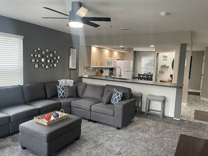 A modern living room with a grey sectional sofa, a small ottoman, and a coffee table. A ceiling fan and recessed lighting are visible.
