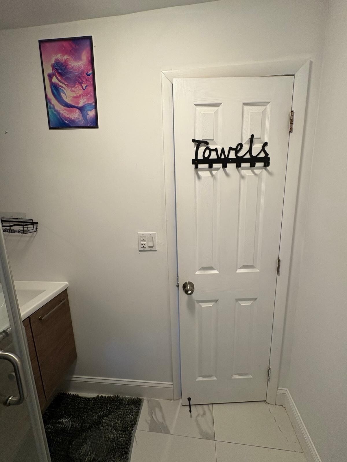 A bathroom with a white door, a towel rack with the word "Towels" on it, a small painting on the wall, and a shower area with a glass door.