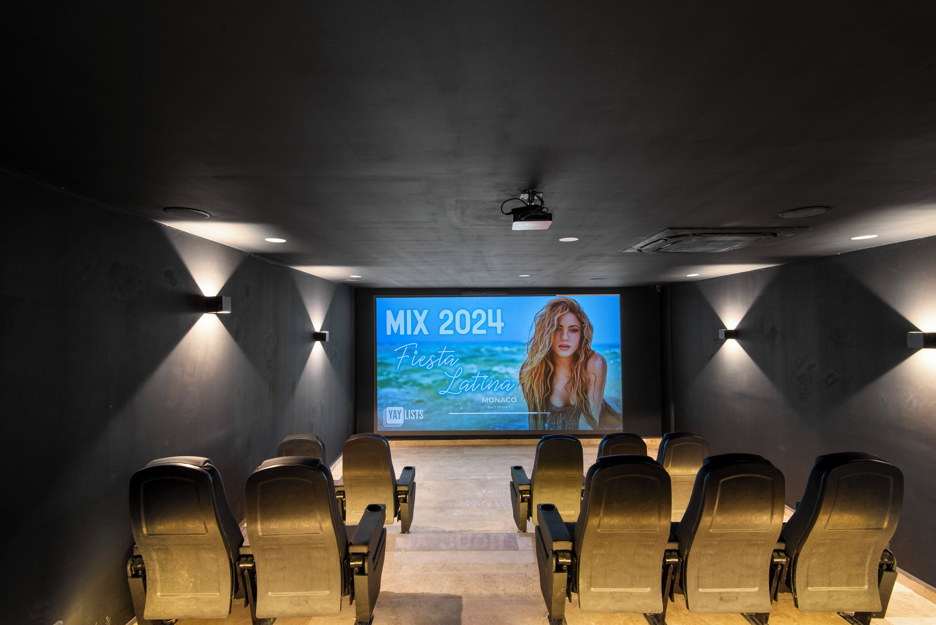 Sala de cinema com fileiras de assentos e uma tela grande exibindo um evento.
