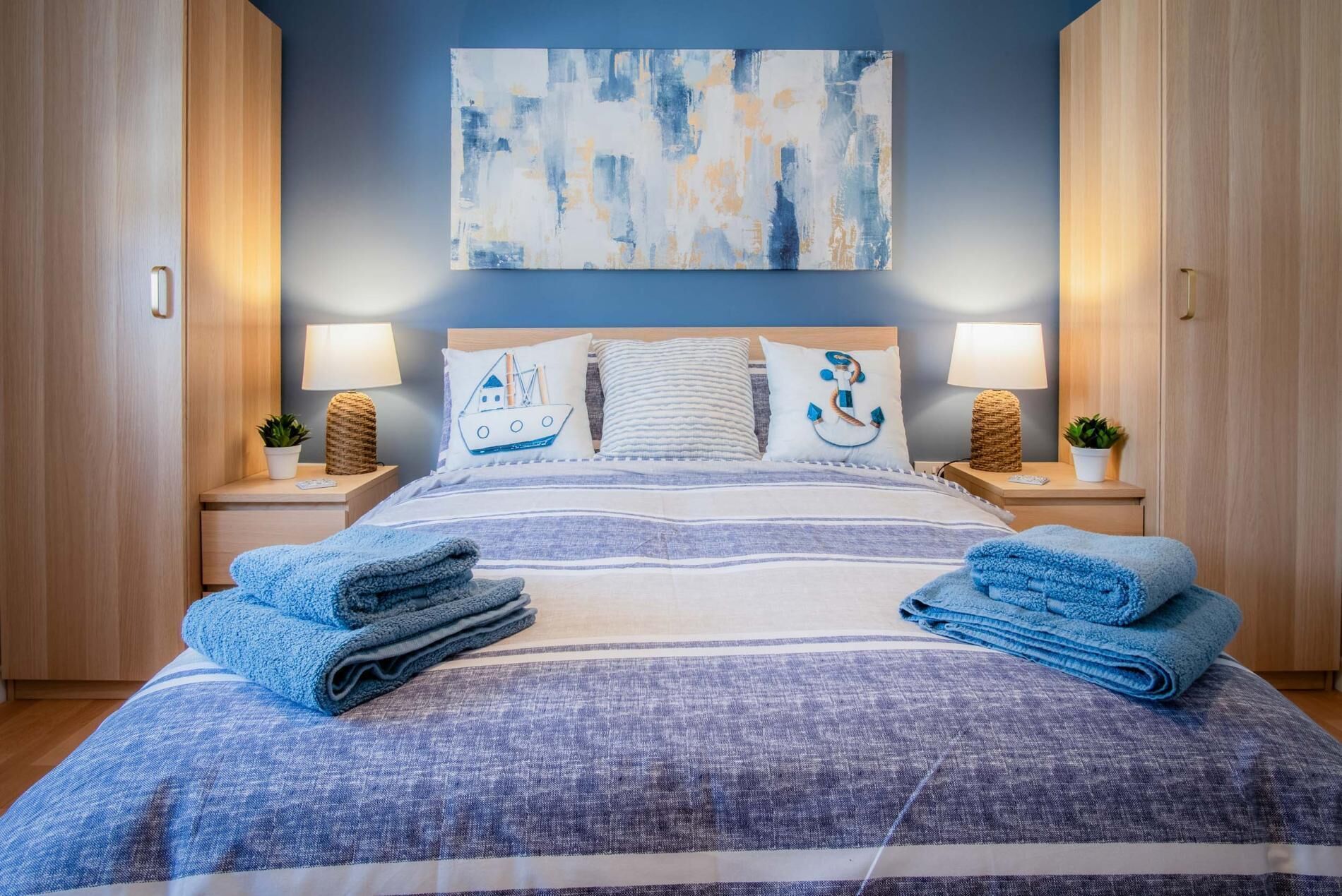Cama bem arrumada com roupa de cama listrada e toalhas azuis, duas luminárias de cabeceira e uma pintura abstrata na parede.