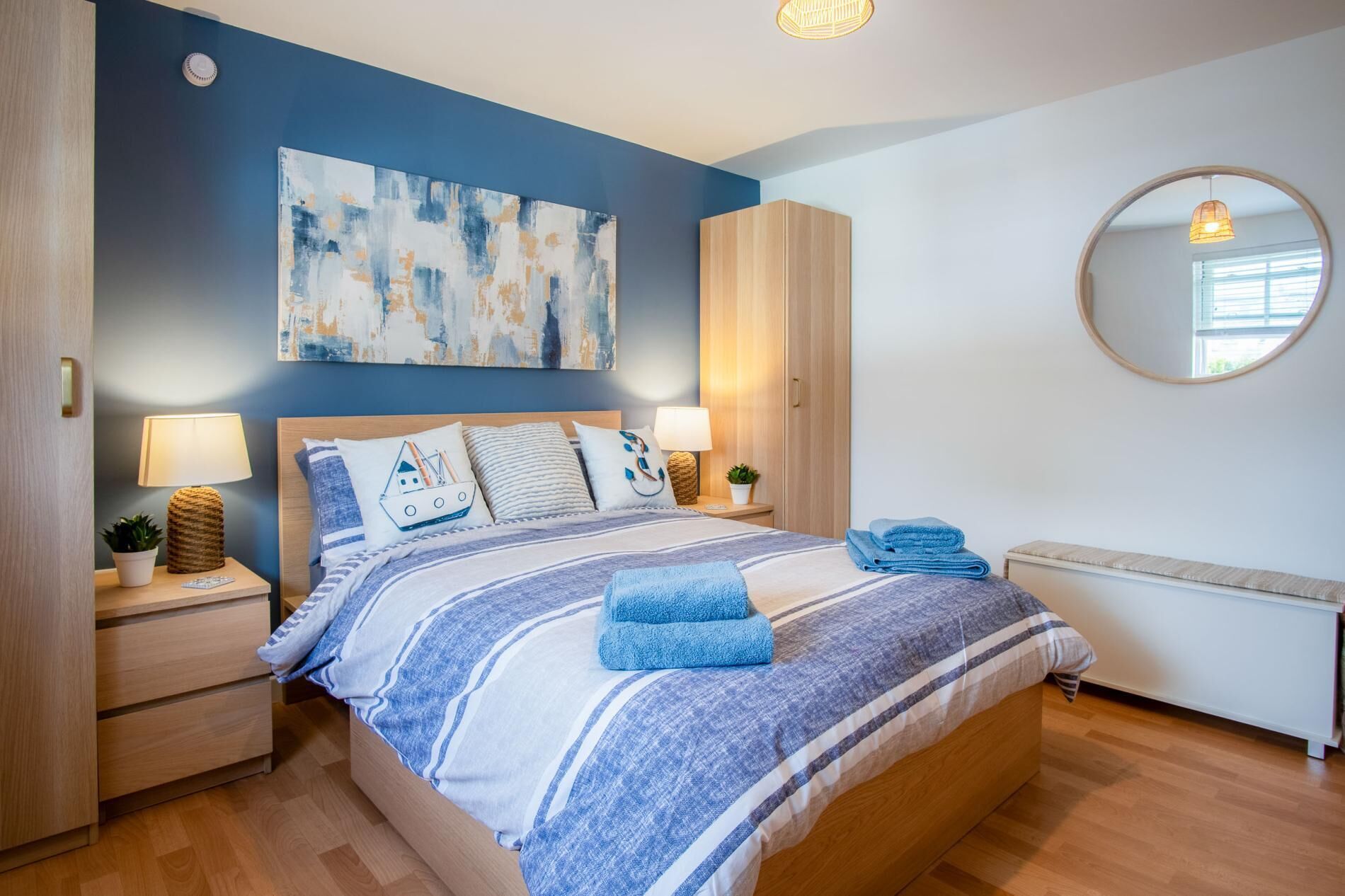 A bedroom with a bed, two bedside tables, a nightstand, a wardrobe, a round mirror, and a wall painting.