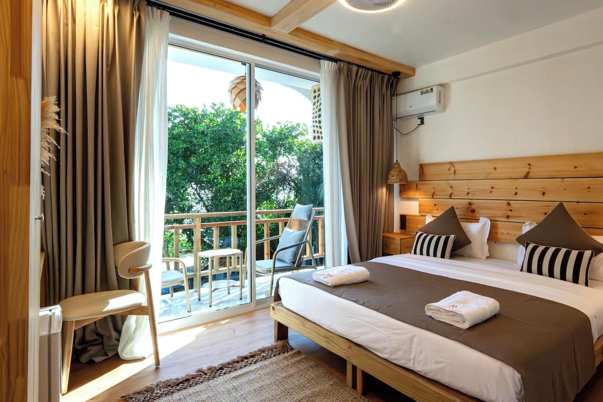 A bedroom with a large bed, a wooden headboard, a balcony with a chair, and a view of trees.
