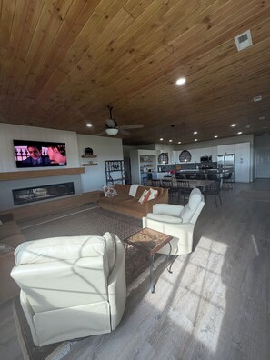 A spacious living area with a wooden ceiling, a flat-screen TV mounted on the wall, a sofa, a coffee table, and a dining area with chairs and a kitchen in the background.