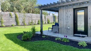 Un patio moderno al aire libre con una pérgola de madera, una zona de estar con una silla y una mesa, y un césped bien cuidado.