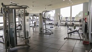 Un gimnasio con diversas máquinas de ejercicio, ventanas amplias y un ventilador de techo.