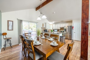 A dining area with a wooden table set for six, surrounded by chairs, a bar stool, and a window with a view of the outdoors.