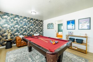 A pool table in a room with a mountain-themed wallpaper, a brown leather sofa, and a bed in the background.
