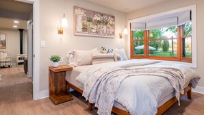 Un dormitorio con una cama grande, una mesita de noche con una planta, un cuadro en la pared y una ventana con vistas a zonas verdes.