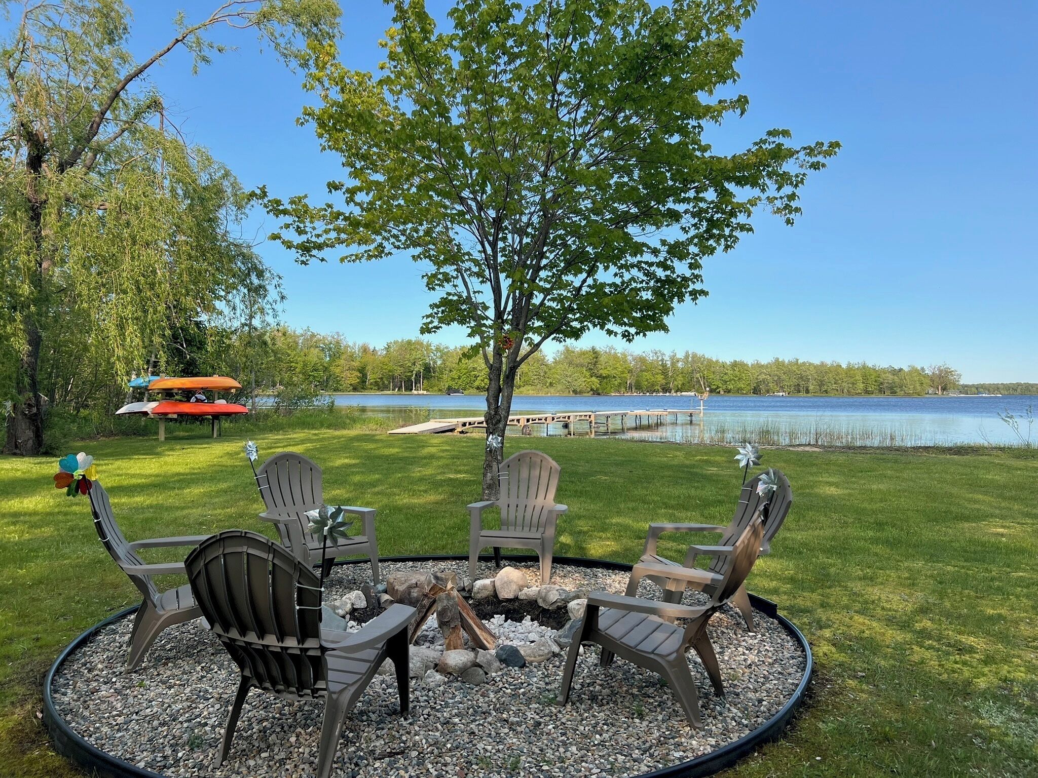 A circular seating area with four Adirondack chairs around a fire pit, a tree in the center, and a lake view in the background.