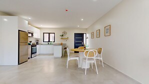 A modern kitchen with white cabinets, a built-in oven, and a refrigerator. There is a dining area with a wooden table and white chairs.