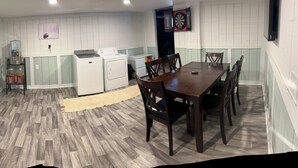 A compact room with a dining table and chairs, a washing machine, a small refrigerator, and a dartboard on the wall.