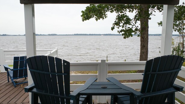 A covered outdoor seating area with two Adirondack chairs and a table, overlooking a body of water. - Riverfront Retreat on Perquimans: Cozy Fire Pit, Golf Minutes Away, Serene Views (Hertford)