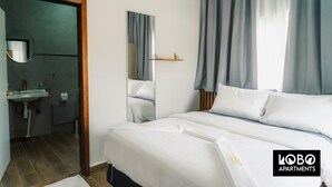Habitación de hotel con cama, mesita de noche, espejo, ventana con cortinas, lavamanos y toallero.