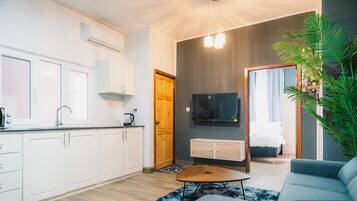 Un apartamento moderno con cocina, sala de estar con sofá y mesa de centro, televisor montado en la pared y una planta grande en maceta.